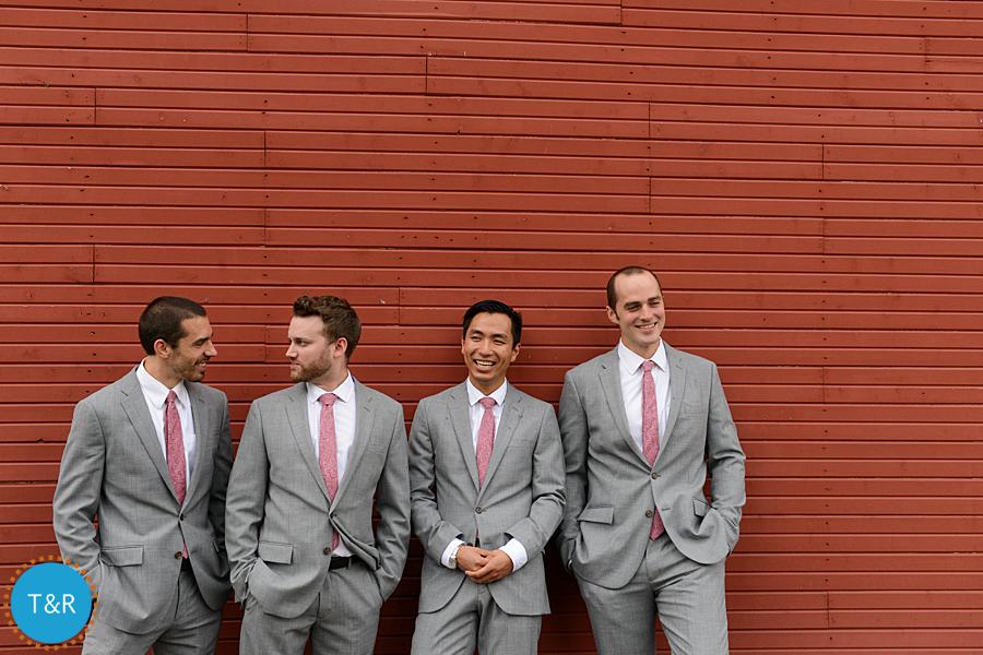 Groom and groomsmen lean against a red barn and laugh together after a Zingerman's Cornman Farms Wedding in Ann Arbor