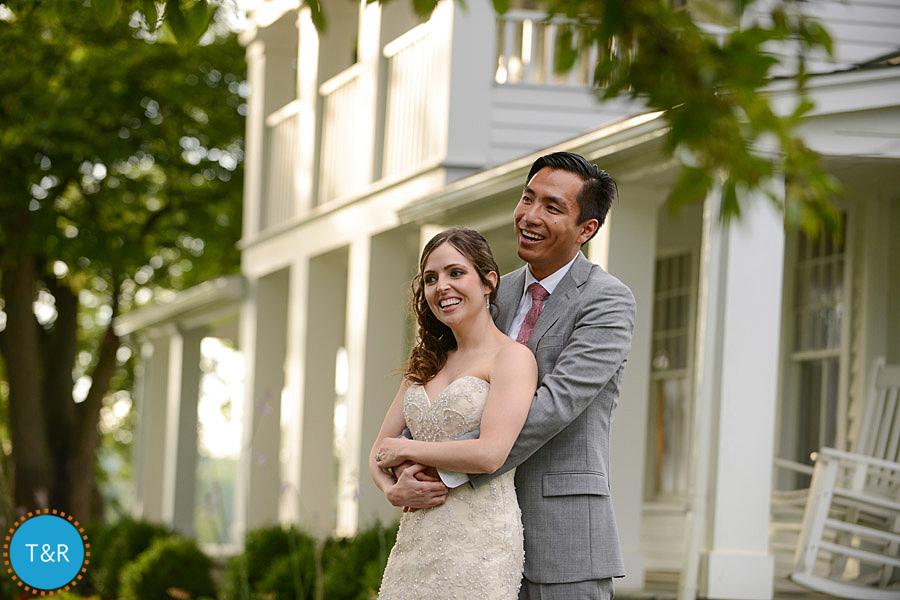 Bride and groom hug and laugh in front of a historic white farmhouse at Zingerman's Cornman Farms Wedding in Ann Arbor