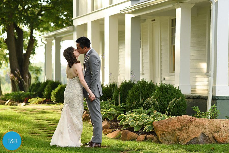 Bride and groom kiss in front of the historic white farmhouse at their Zingerman's Cornman Farms wedding reception in Ann Arbor, MI