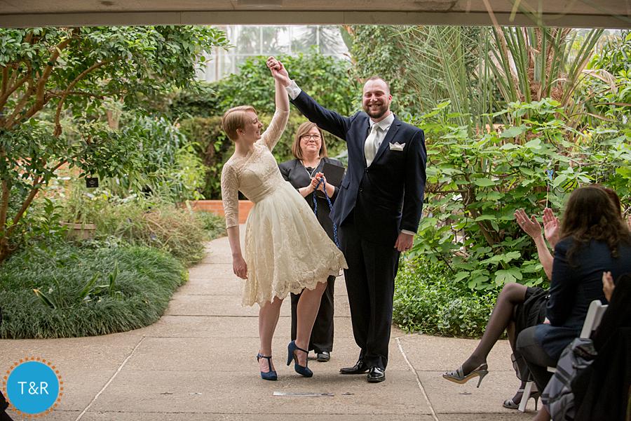Bride and groom raise their clasped hands surrounded by tropical plants after their Matthaei Botanical Gardens wedding ceremony