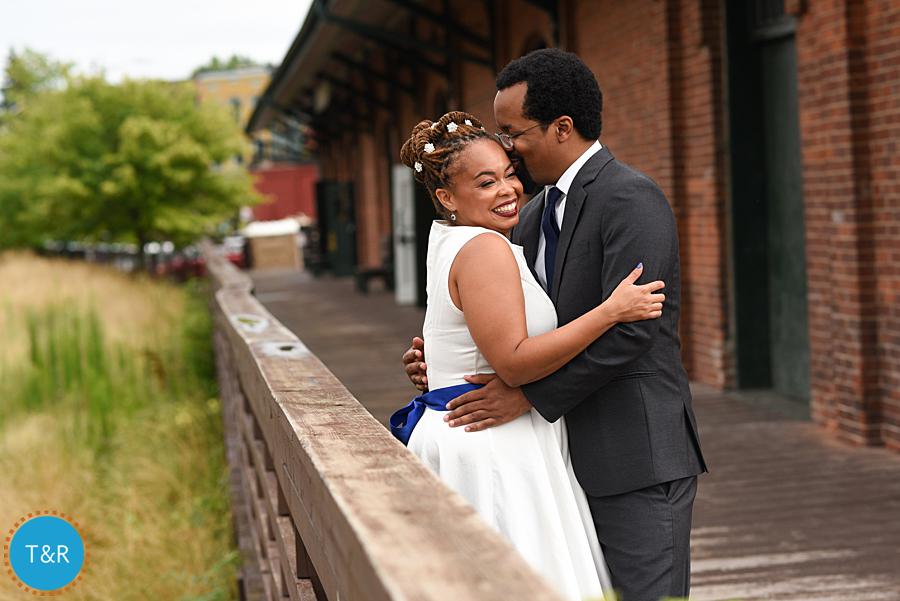 Bride and groom smile and kiss while leaning on a rail outside a train depot after their wedding in Ypsilanti's Depot Town