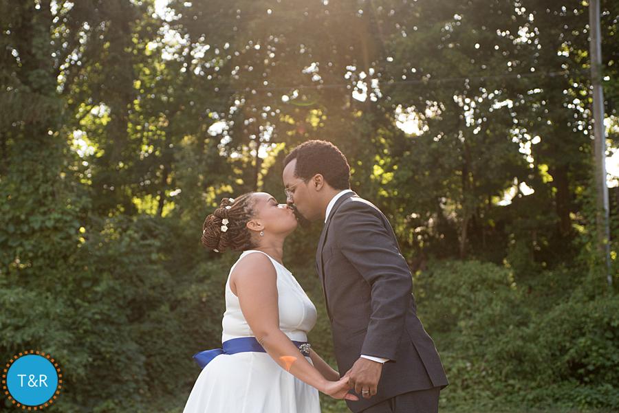 Bride and groom kiss while holding hands against a backdrop of trees and sunshine during their Ypsilanti wedding